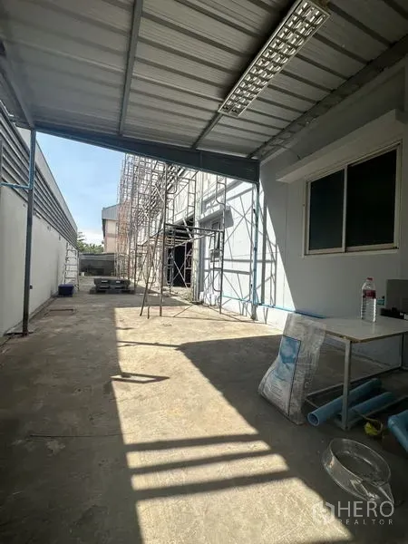 Factory for rent or sale in Bang Bua Thong, Nonthaburi - Side service yard under metal roof with scaffolding and storage area along the factory wall.