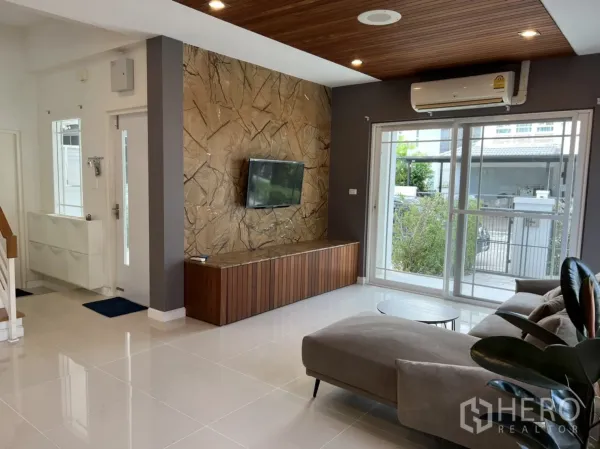 Detached House for rent in Bang Kapi, Bangkok - Bright living room with sliding glass doors, air‑conditioner, stone feature wall with TV, and an L‑shaped sofa.