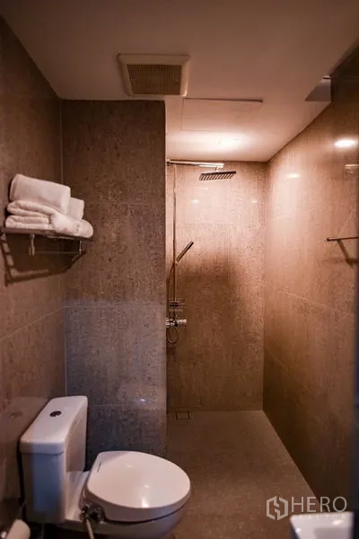 Condominium for rent in Khlong Toei, Bangkok - Second bathroom with rain shower, toilet, towel shelf, and warm stone tiles.