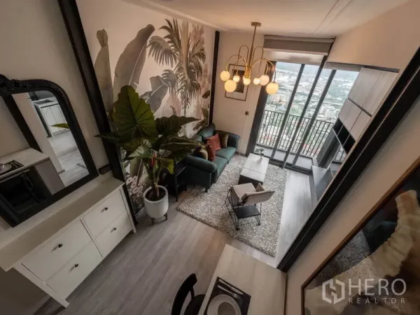 Condominium for rent or sale in Phra Khanong, Bangkok - Stylish living room with tropical mural, chandelier, green sofa and balcony city view through floor-to-ceiling glass.