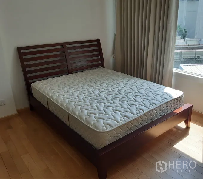 Condominium for rent in Khlong Toei, Bangkok - Sunlit bedroom with wooden bed frame and mattress beside a large window with curtains.