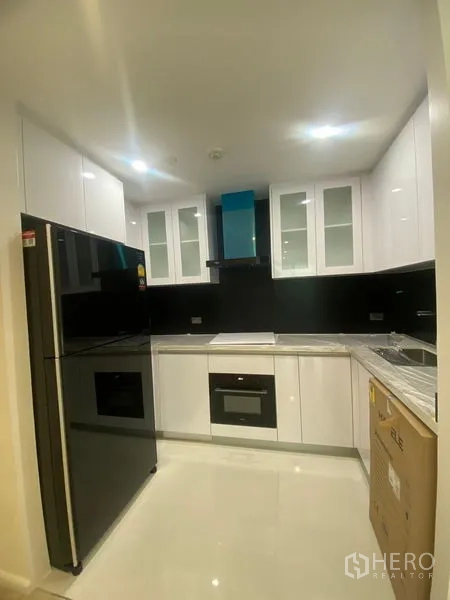 Apartment for rent in Watthana, Bangkok - Compact L‑shaped kitchen with glossy white cabinets, black backsplash, built‑in oven and large refrigerator.