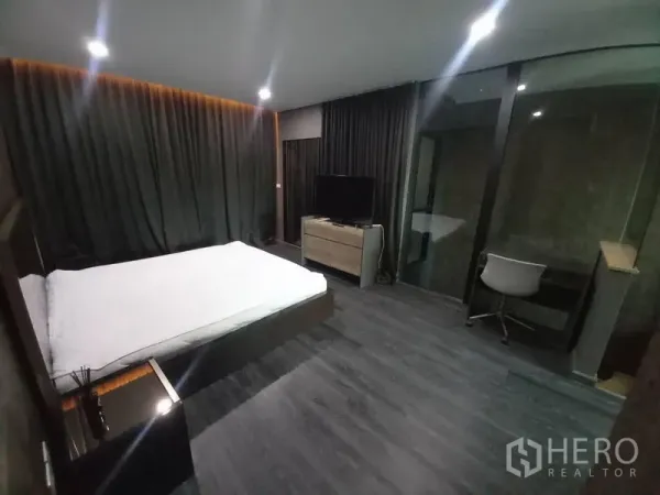 Townhouse for rent in Suan Luang, Bangkok - Spacious master bedroom with dark flooring, blackout curtains, TV cabinet, glass-walled workspace and ensuite access.