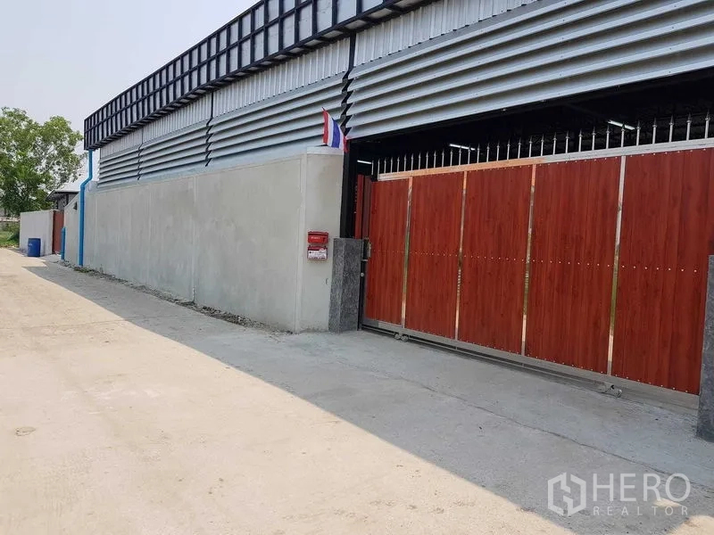 Warehouse for sale in Lam Luk Ka, Pathum Thani - Street-side view of a secure warehouse with a tall red sliding gate and concrete perimeter wall in Lam Luk Ka.
