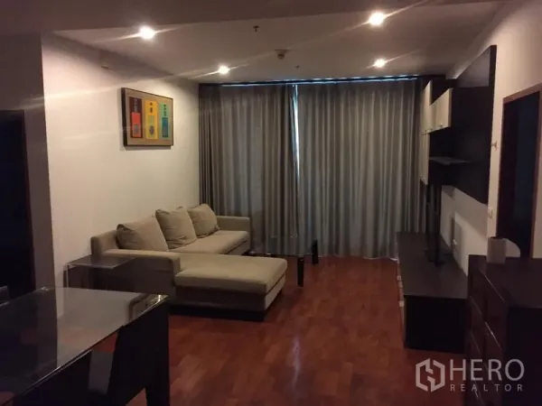 Condominium for rent or sale in Khlong Toei, Bangkok - Cozy living room with L-shaped sofa, wood flooring, media wall and full-length blackout curtains.