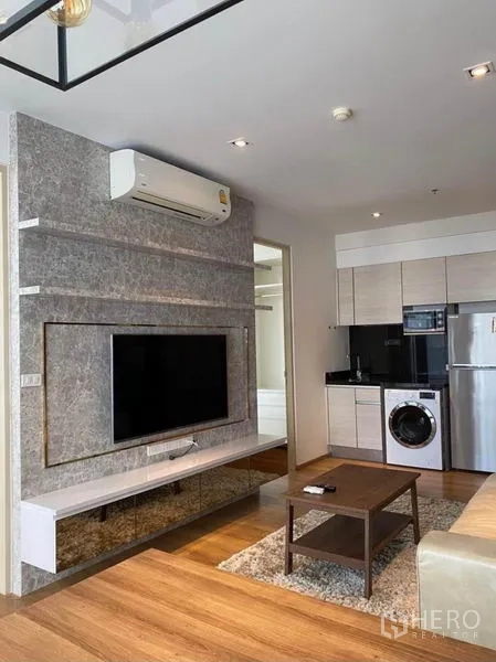 Condominium for rent in Khlong Toei, Bangkok - Open-plan living with wall-mounted TV and compact kitchen featuring white cabinetry, fridge, and washing machine.