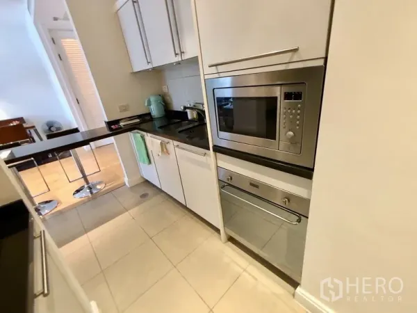 Condominium for rent in Pathum Wan, Bangkok - Compact modern kitchen with white cabinetry, black countertops, built‑in microwave and oven, and a breakfast bar with stools.