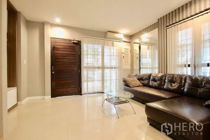 Townhouse for sale in Suan Luang, Bangkok - Living room with wooden front door, large windows, and sectional sofa.