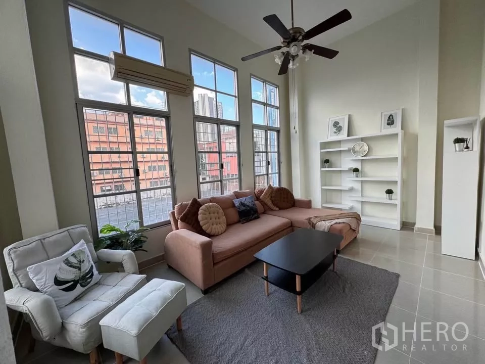 Townhouse for rent in Bang Kho Laem, Bangkok - Living room with floor-to-ceiling windows, ceiling fan, sofa set and white shelving.