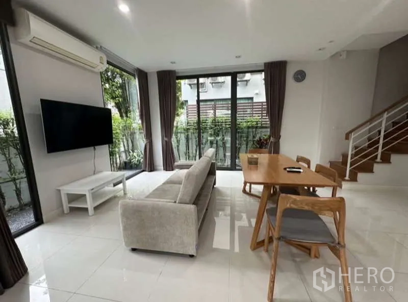 Townhouse for rent in Lat Phrao, Bangkok - Open-plan living and dining area with sofa, wall-mounted TV, dining set and floor-to-ceiling windows.
