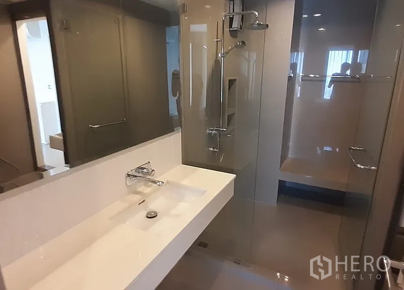 Condominium for rent in Khlong Toei, Bangkok - Modern bathroom with long vanity, large mirror, and glass-enclosed shower.