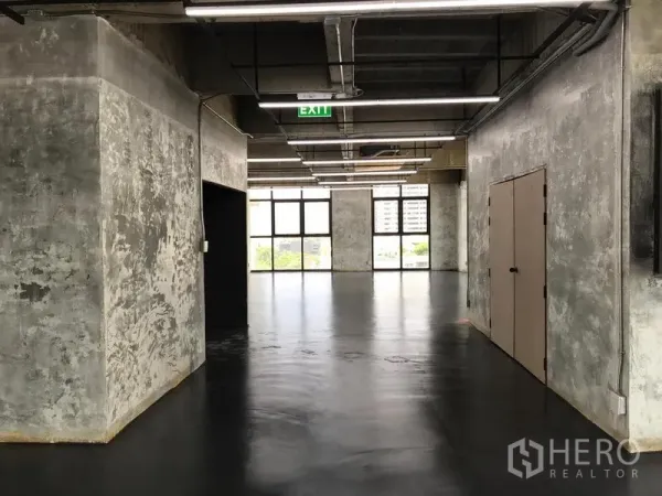 Office Space for rent in Khlong Toei, Bangkok - Hallway view of raw-finished office with textured concrete walls, linear lights, exit sign, and wide windows overlooking the city.