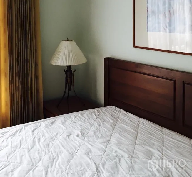 Condominium for rent in Watthana, Bangkok - Bedroom corner featuring a wooden headboard, bedside lamp, long curtains and framed wall art.