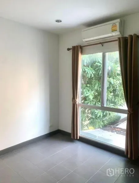Townhouse for rent in Suan Luang, Bangkok - Ground-floor room with large window, brown curtains and split air conditioner facing greenery.