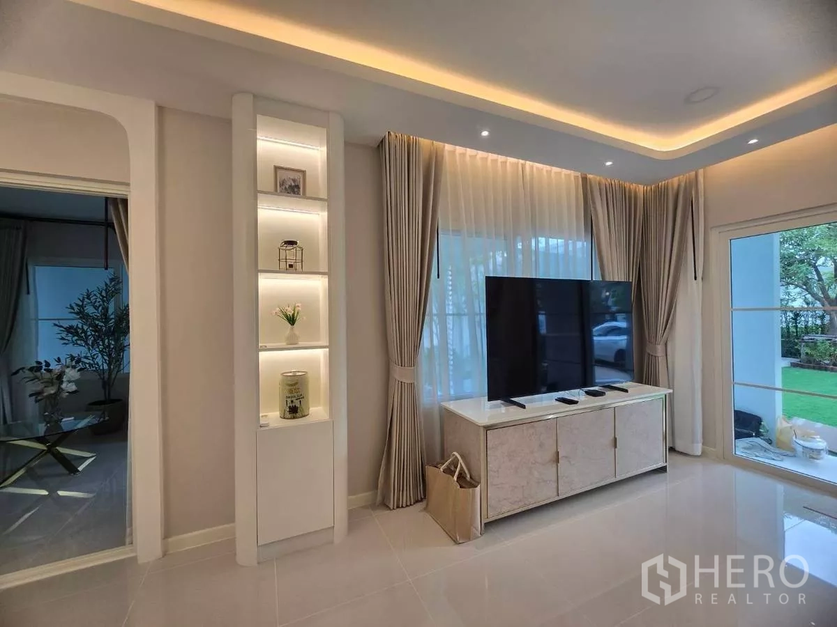 独立式住宅 出租在Bang Phli, Samut Prakan - 客厅配电视柜、带灯展示架与落地窗帘。
