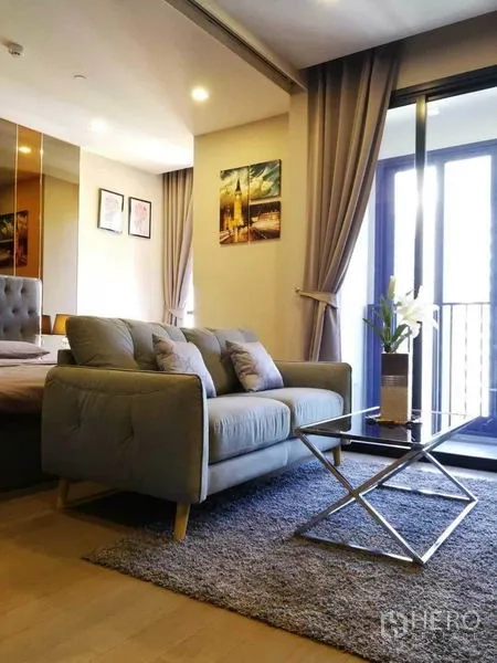 Condominium for rent in Watthana, Bangkok - Bright living area with gray sofa, glass coffee table, rug and sliding doors to a balcony.