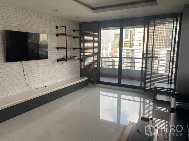 Condominium for rent in Khlong Toei, Bangkok - Living room with balcony access, white brick feature wall, mounted TV, and skyline view.