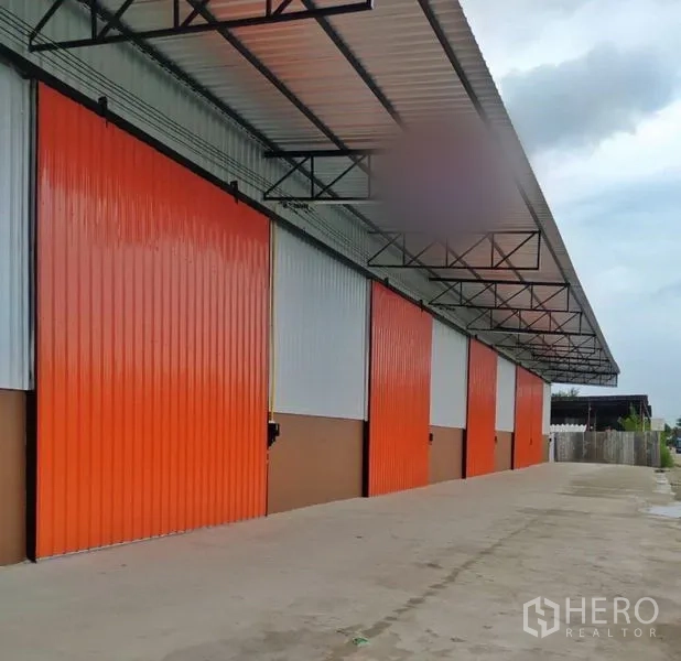 货栈 出租在Khlong Luang, Pathum Thani - 一排现代仓库，橙色推拉门，宽阔混凝土车道及带顶装卸区。
