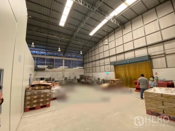 Warehouse for rent in Bueng Kum, Bangkok - High-ceiling warehouse interior with steel trusses, louvered windows, pallets, and a yellow PVC strip door.
