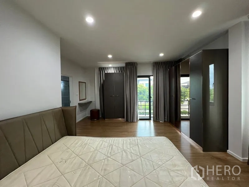 Detached House for rent in Saphan Sung, Bangkok - Spacious master bedroom with wood flooring, large bed, built‑in wardrobes and a balcony door with curtains.