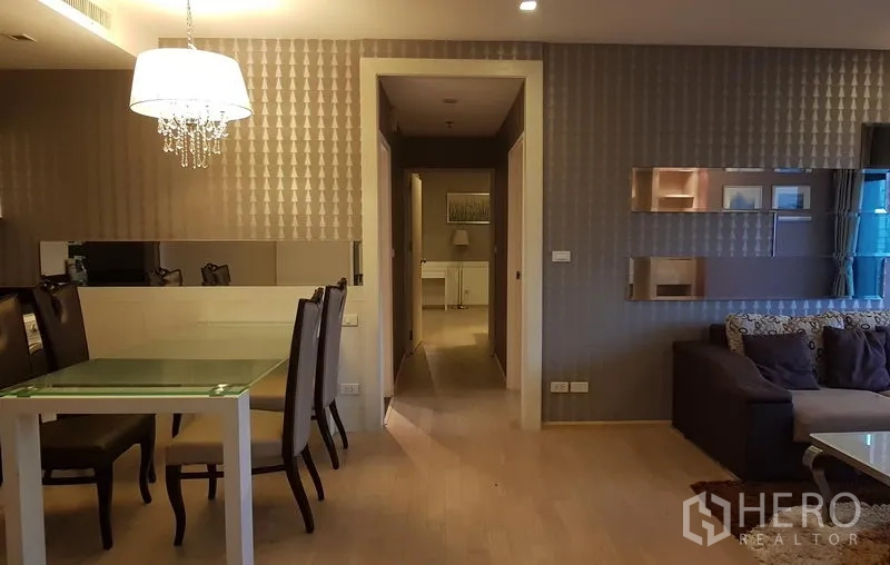 Condominium for rent in Khlong Toei, Bangkok - Dining area with glass table and chandelier beside a mirrored feature wall leading to the bedroom hallway.