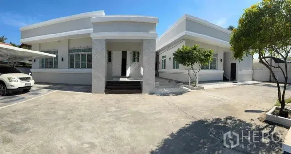 独立式住宅 出租在Samut Prakan - 正立面全景，宽阔车道、车棚与两栋单层建筑。
