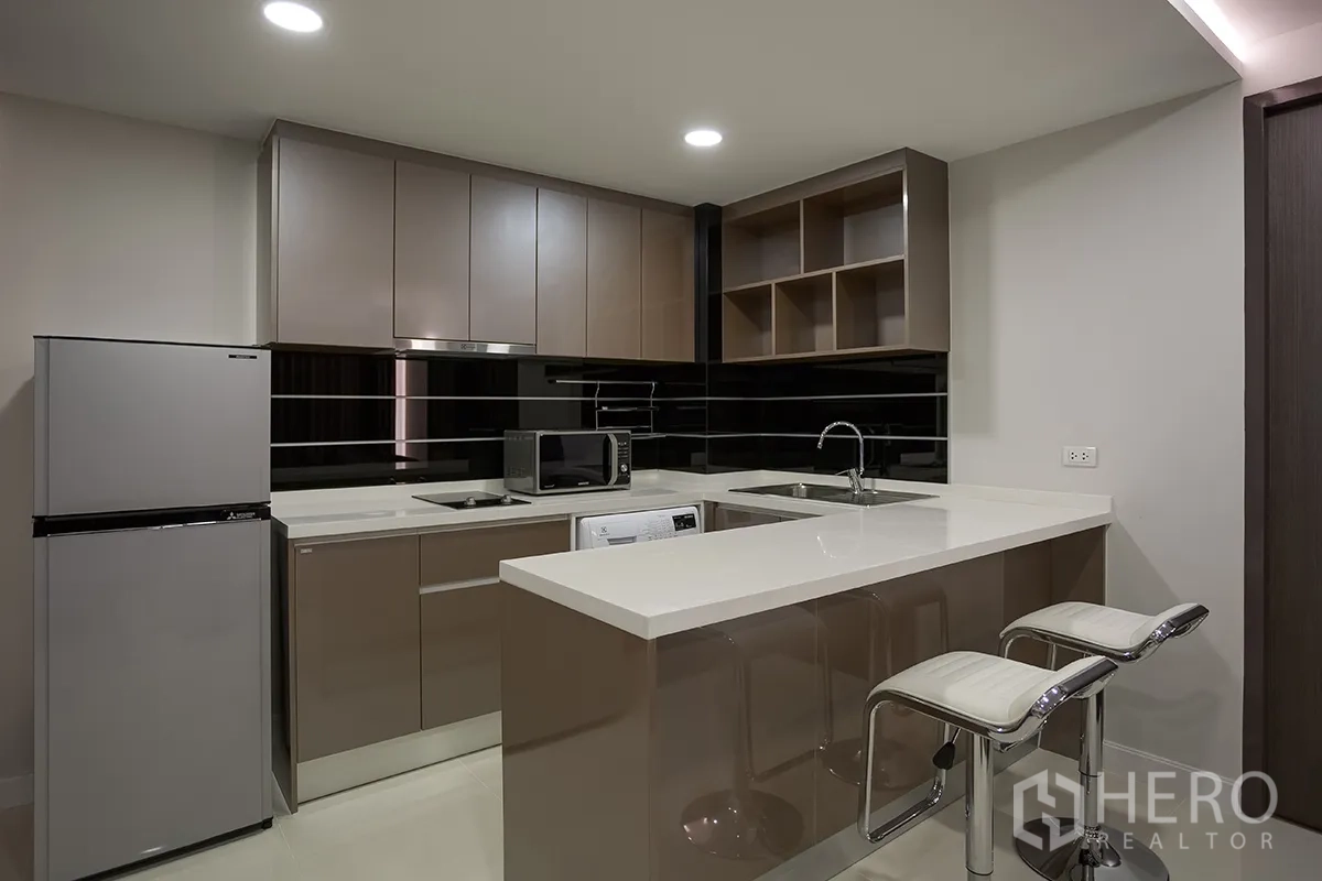 Condominium for rent in Khlong Toei, Bangkok - Modern kitchen with breakfast bar, glossy cabinets, fridge, microwave, sink, and washing machine.