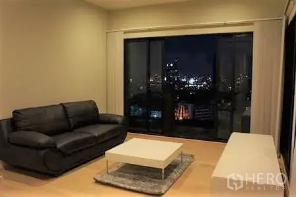 Condominium for rent in Watthana, Bangkok - Living room with black leather sofa, low coffee table and floor‑to‑ceiling sliding glass doors overlooking the Bangkok skyline at night.