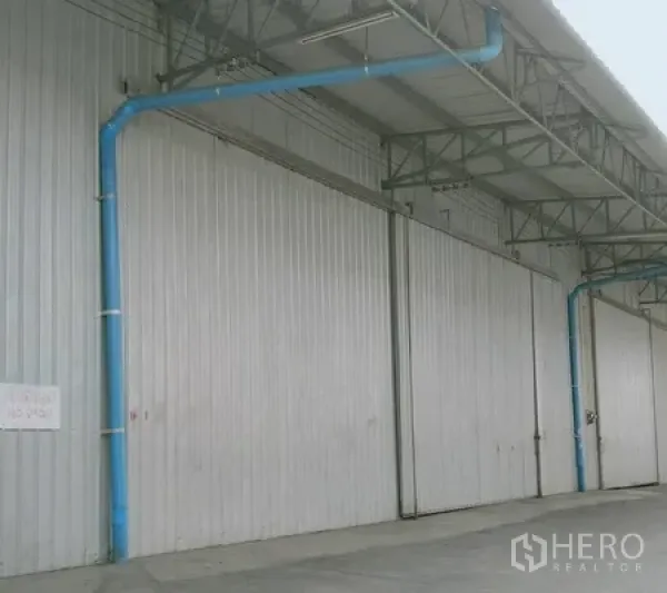 Warehouse for rent in Khlong Luang, Pathum Thani - Exterior of a steel-clad warehouse with large sliding metal doors and covered loading bay with blue piping.