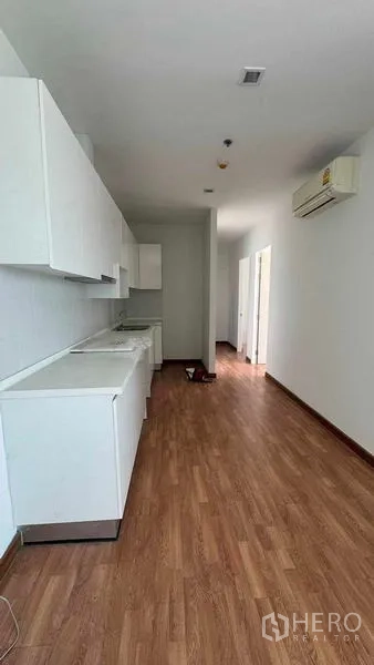 公寓 出租在Bang Na, Bangkok - 长廊式厨房，白色橱柜与操作台，水槽、木地板及空调沿墙布置。