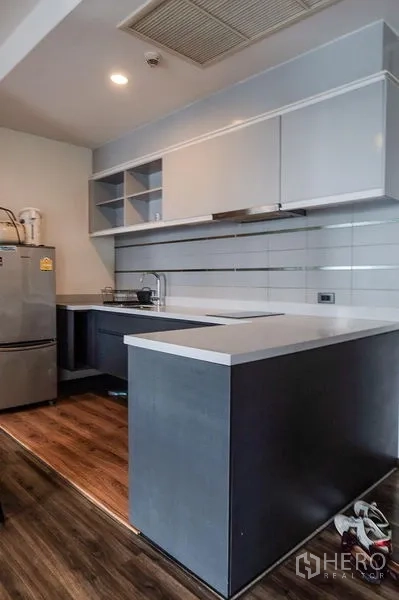 Condominium for rent in Khlong Toei, Bangkok - L-shaped kitchen with upper cabinets, electric cooktop and stainless refrigerator.