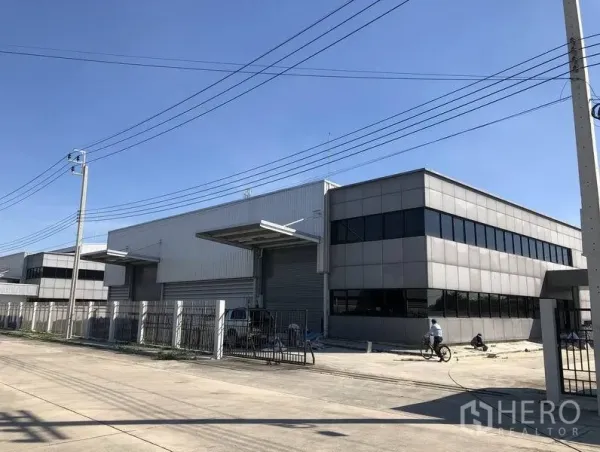 Factory for rent or sale in Bang Sao Thong, Samut Prakan - Modern factory exterior with grey cladding, covered loading bays, large roller shutters and a fenced frontage on a wide road.