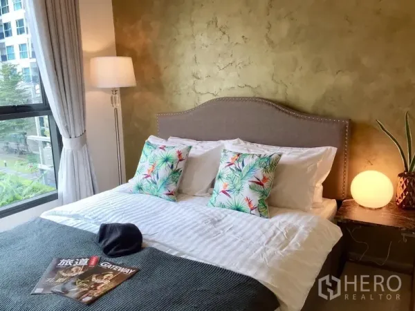 Condominium for rent in Khlong Toei, Bangkok - Bright bedroom with large window, textured gold accent wall, double bed with patterned cushions and bedside lamp.