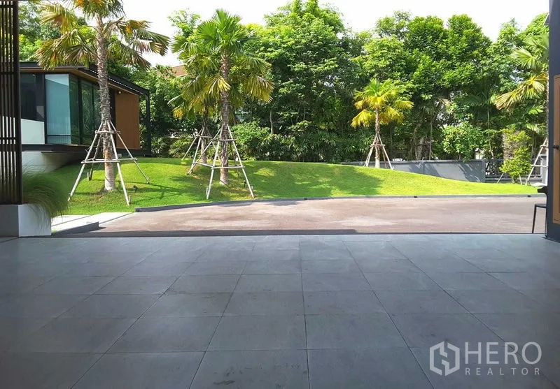 Detached House for rent or sale in Lam Luk Ka, Pathum Thani - Covered terrace opening to a landscaped driveway with palm trees and raised grassy mounds.