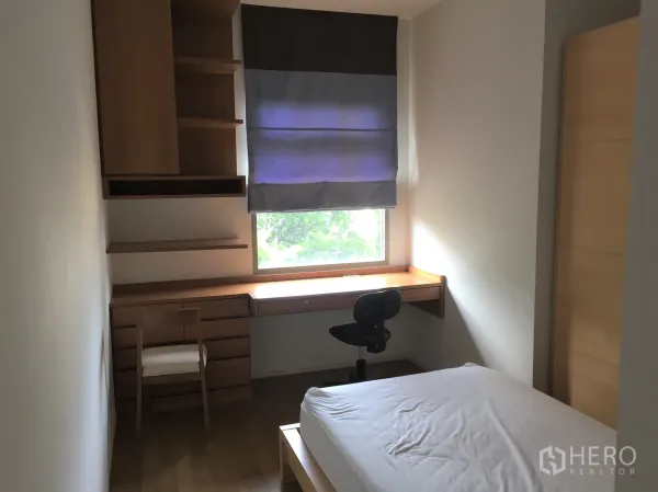 公寓 出租在Watthana, Bangkok - 次卧带单人床、内置书桌与搁架，窗外绿意，配罗马帘。