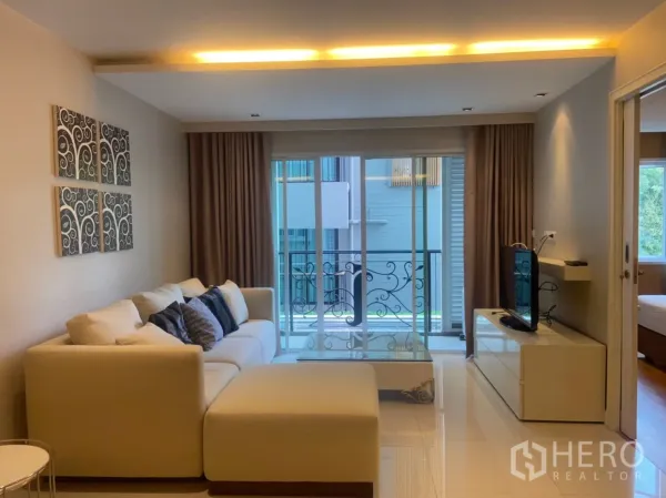 คอนโด ให้เช่าในWatthana, Bangkok - ห้องนั่งเล่นอบอุ่น โซฟารูปตัวแอล เคาน์เตอร์ทีวี และประตูกระจกสไลด์ออกสู่ระเบียงราวเหล็กดัด