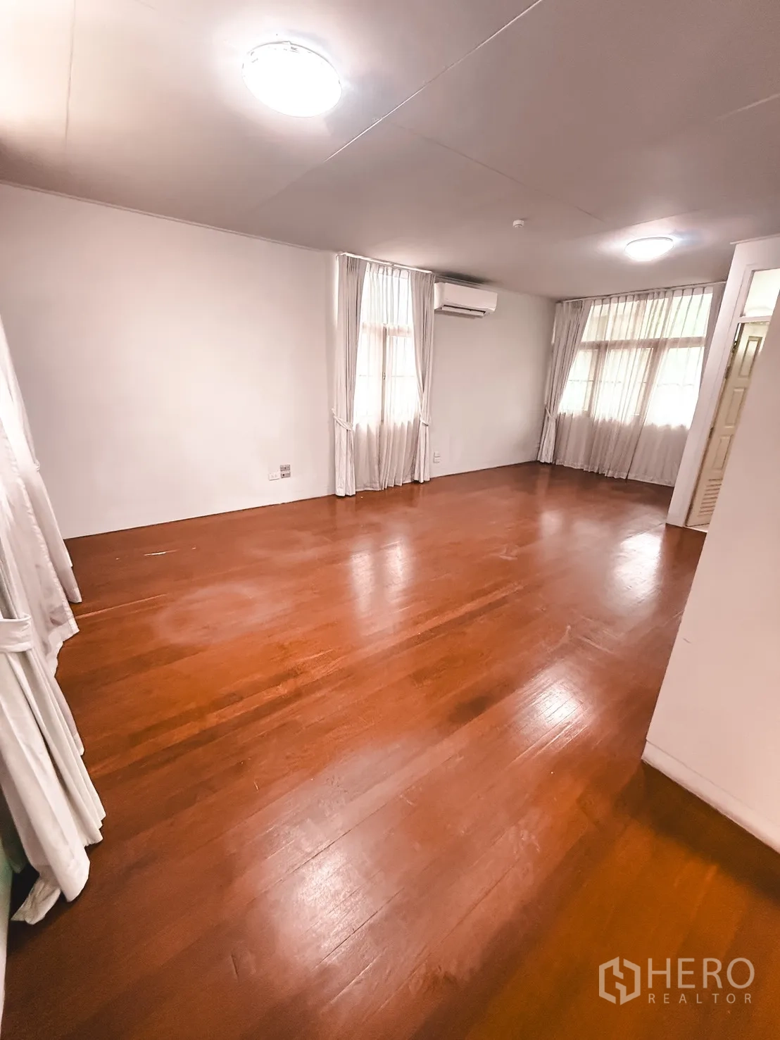 Home Office for rent in Watthana, Bangkok - Spacious room with hardwood floors, two curtained windows, and a wall-mounted air conditioner.
