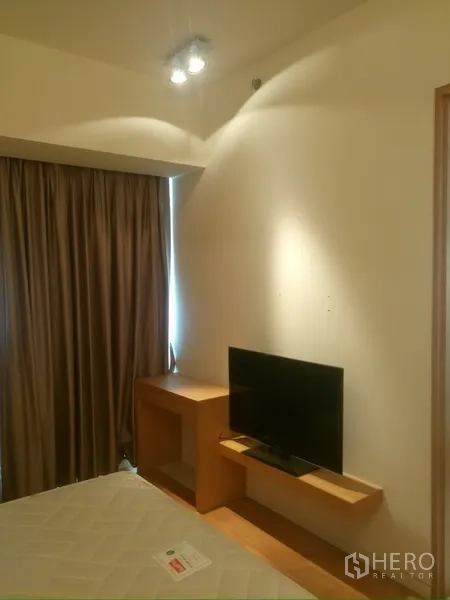 Condominium for rent in Sathon, Bangkok - Bedroom corner with TV on floating shelf and a desk beside a curtained window.