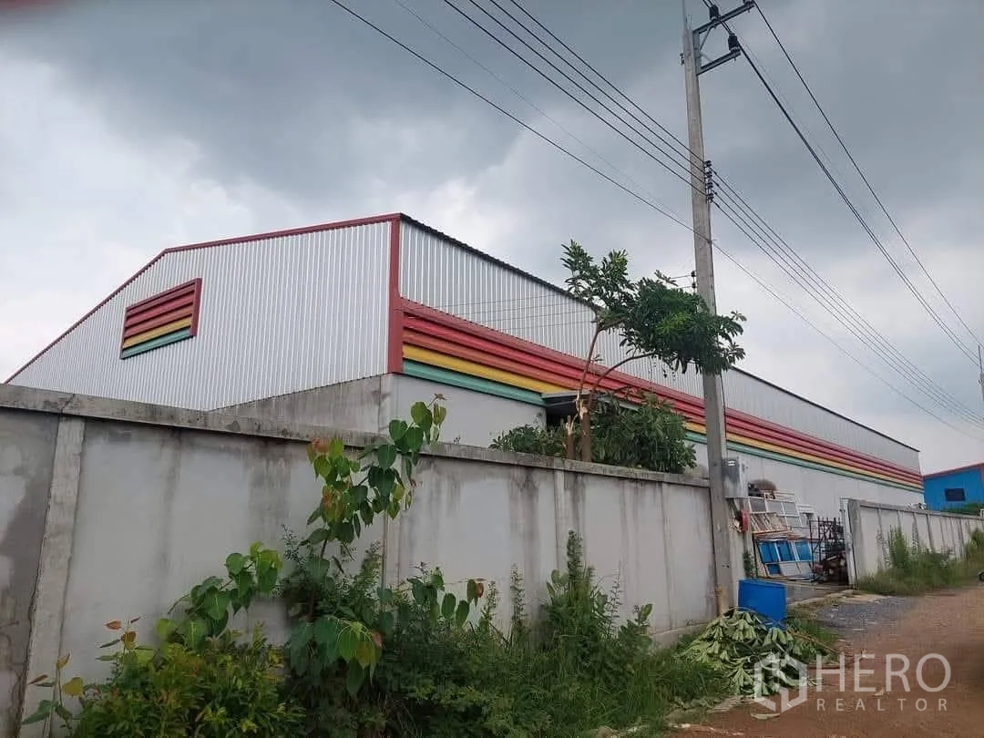 仓库 for sale in Lam Luk Ka, Pathum Thani - 仓库外观斜角视图，凸显高坡屋顶、围墙与路边植被。