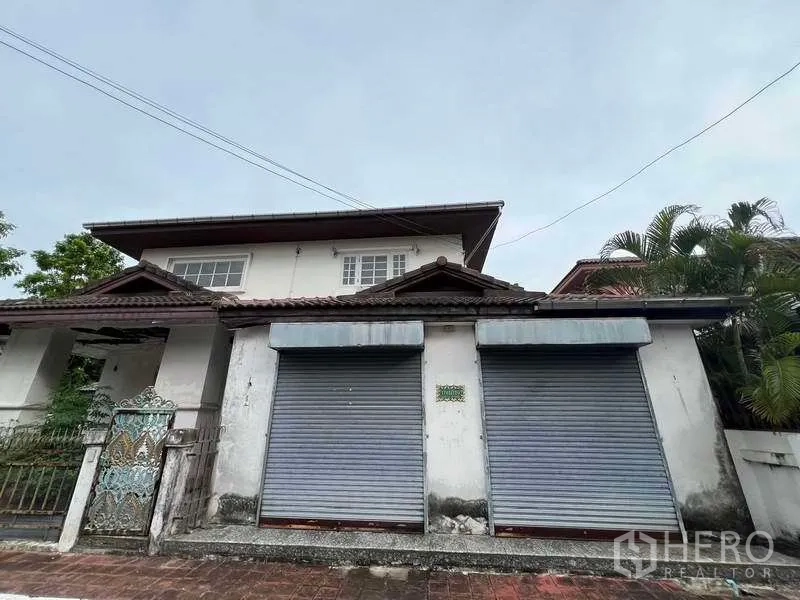 独立式住宅 出售在Phra Khanong, Bangkok - 正面局部特写，展示双卷帘门、屋檐瓷瓦与墙侧热带植物。