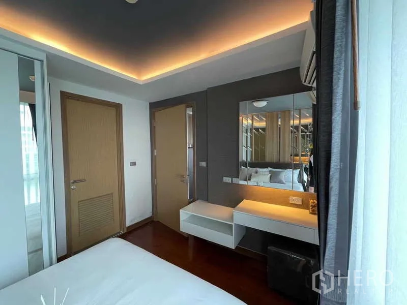 Condominium for sale in Watthana, Bangkok - Bedroom angle showing floating dressing table, large mirror wall and warm cove lighting by the window.