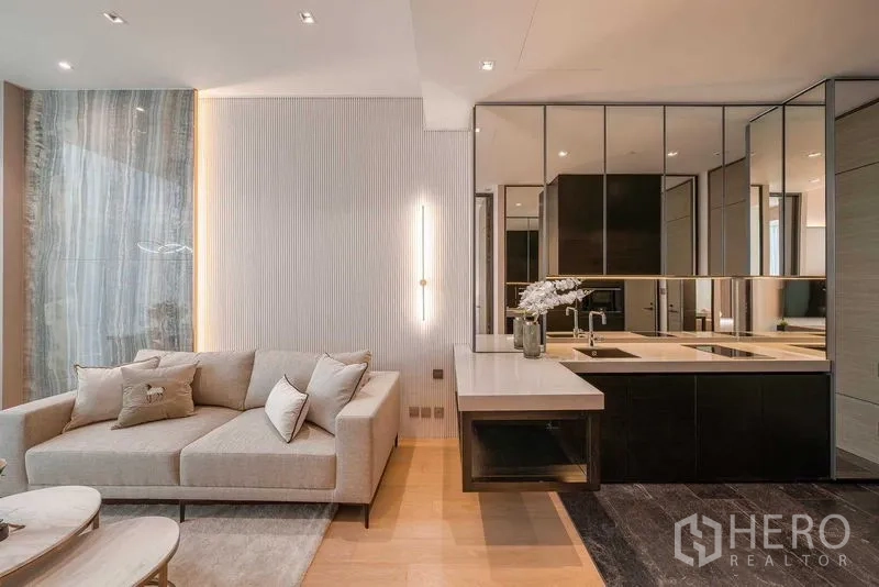 Condominium for rent or sale in Pathum Wan, Bangkok - Contemporary living room with beige sofa, compact kitchen island and mirrored cabinetry.