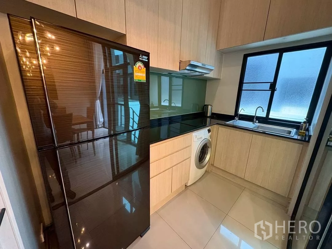 Townhouse for rent in Suan Luang, Bangkok - Compact fitted kitchen with black refrigerator, light wood cabinetry, sink and washing machine.