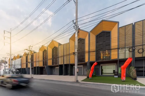 Warehouse for rent in Mueang Samut Sakhon, Samut Sakhon - Row of modern loft-style warehouses with gabled facades facing a main road, wide driveways and red air dancers at the entrance at dusk.