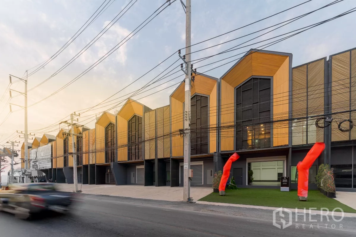 Warehouse for rent in Mueang Samut Sakhon, Samut Sakhon - Row of modern loft-style warehouses with gabled facades facing a main road, wide driveways and red air dancers at the entrance at dusk.