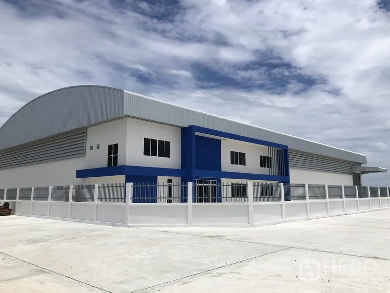 Warehouse for rent in Samut Prakan - Modern white-and-blue warehouse exterior with curved metal roof and perimeter fence under a bright sky.
