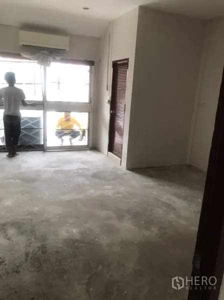 Home Office for rent in Khlong Toei, Bangkok - Bright room with air conditioner, large window with grille, unfinished floor, and a door to a side room.