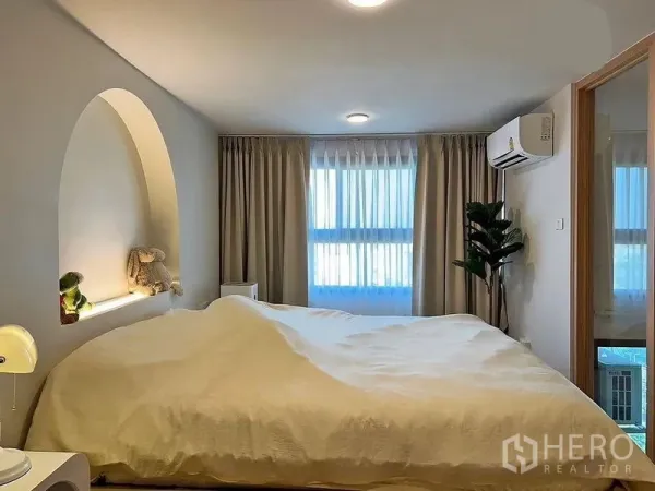 Condominium for rent in Huai Khwang, Bangkok - Bright bedroom with curved alcove headboard shelf, large windows, curtains and wall-mounted air conditioner.
