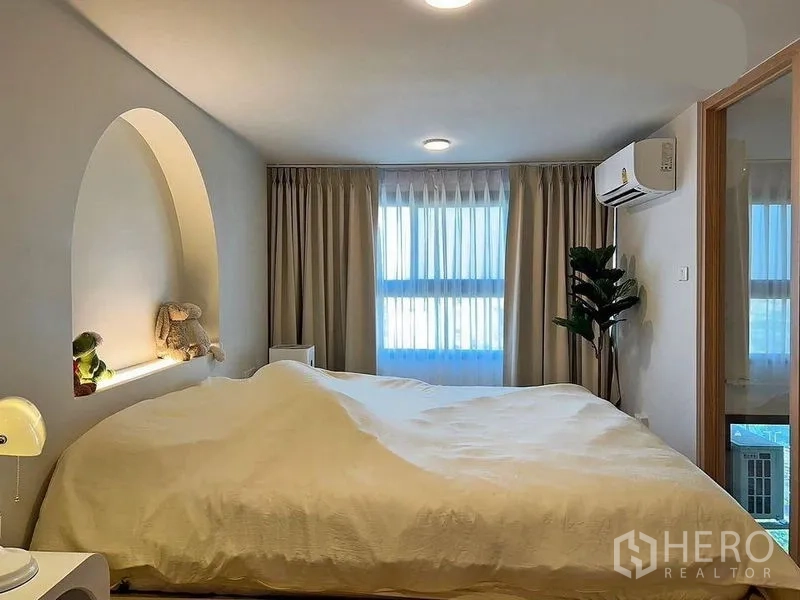 Condominium for rent in Huai Khwang, Bangkok - Bright bedroom with curved alcove headboard shelf, large windows, curtains and wall-mounted air conditioner.