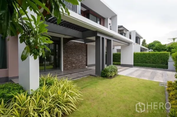Detached House for rent in Prawet, Bangkok - Side facade with stone feature wall, neat lawn and paved parking area.